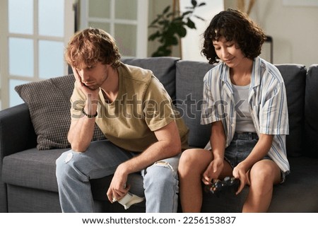 Upset guy with gamepad keeping hand on cheek and feeling sad while sitting on couch next to his girlfriend who won video game Royalty-Free Stock Photo #2256153837