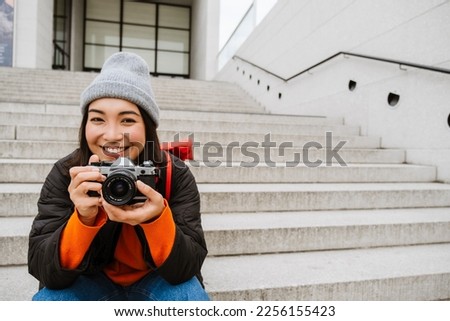 Beautiful young smiling asian woman in warm clothes taking pictures with vintage camera while sitting on stairs at city street