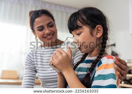 Caucasian little kid holding a cup of milk and drinking with mother. Attractive mom teach and support young girl daughter take care of her body, sipping a milk after wake up for health care in house.
