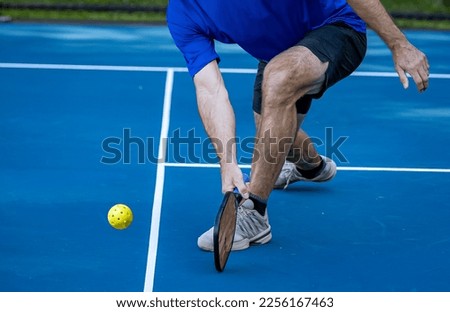 Male pickleball player digs deep to scoop up the ball off the co Royalty-Free Stock Photo #2256167463