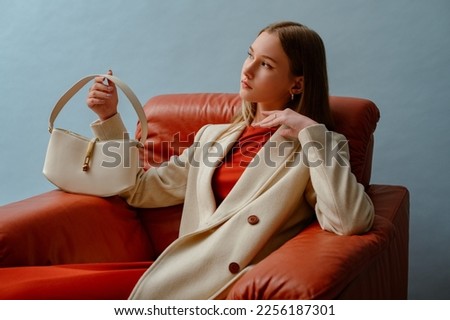 Fashionable confident woman wearing elegant white woolen coat,  holding stylish leather bag, sitting in armchair, posing on blue background. Copy, empty space for text Royalty-Free Stock Photo #2256187301