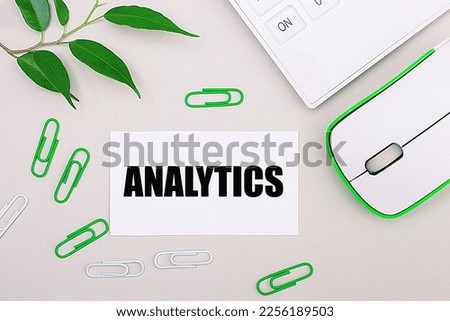 On a light background, a white calculator, a computer mouse, green paper clips, a green plant and a white blank sheet with the text ANALYTICS. Flat lay.