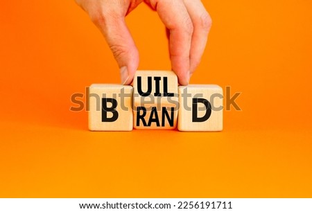 Build your brand symbol. Concept word Build brand on wooden cubes. Beautiful orange table orange background. Businessman hand. Business and build your brand concept. Copy space. Royalty-Free Stock Photo #2256191711