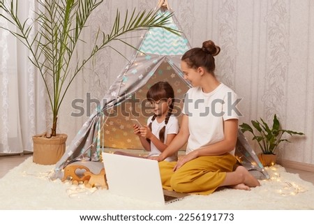 Image of Caucasian dark haired woman sitting on floor near children's teepee with her daughter, little girl using mobile phone, watching cartoons, family use laptop.