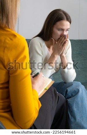 Young sad woman is sitting on the couch, holding her hands to her face at a psychotherapist's appointment. Psychologist with a client. Photo from the psychologist's back. Vertical photo