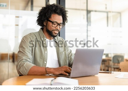 Focused curly handsome man using laptop computer in contemporary coworking space, freelance programmer in glasses coding sitting at the office desk, creativity designer or architect typing Royalty-Free Stock Photo #2256203633