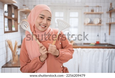 Young Asian Muslim woman housewife wearing Hijab cooking and holding pan and spatula on kitchen background. Royalty-Free Stock Photo #2256209479