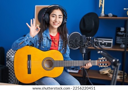Young teenager girl playing classic guitar at music studio doing ok sign with fingers, smiling friendly gesturing excellent symbol 