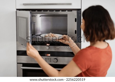 Young woman putting plate with food in the microwave oven in modern kitchen interior, hungry millennial female warming rice with vegetables dish for lunch or dinner at home, closeup shot Royalty-Free Stock Photo #2256220519