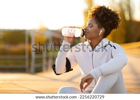 Tired Black Female Jogger Drinking Water While Relaxing After Running Outdoors, Young African American Woman Listening Music In Wireless Headphones And Enjoying Refreshing Drink, Copy Space Royalty-Free Stock Photo #2256220729