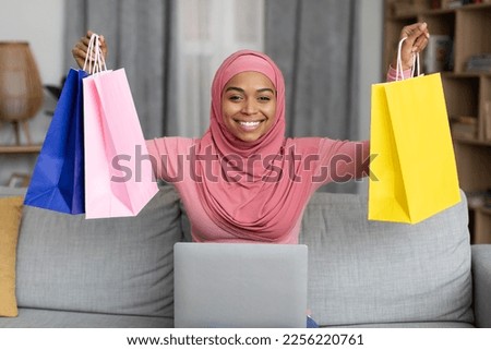 Excited black lady in hijab sitting on sofa with laptop and lifting gift bags above her head at home. Charming african american muslim woman purchasing presents on web, happy to get big discount