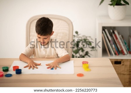 Busy smiling small european boy with colorful hands palms enjoys drawing at table in white room interior. Schooling, entertainment and fun, education, dreams, art and fantasy, childhood at home