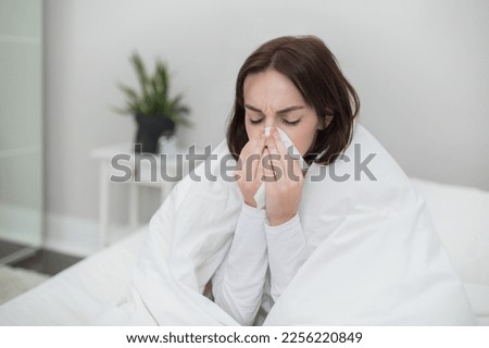 Sixk unhappy millennial brunette lady in pajamas sitting in bed covered in warm blanket at home, sneezing, using napkin, suffering from cold, flu or coronavirus concept, copy space, closeup Royalty-Free Stock Photo #2256220849