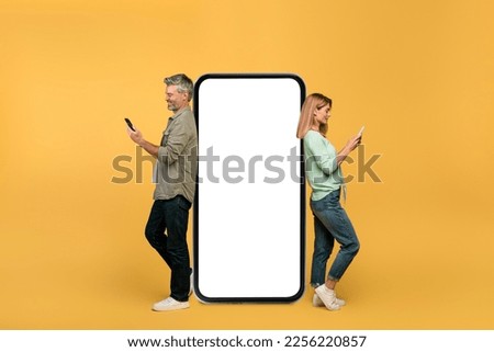 Middle aged man and woman using smartphones, leaning on large phone with empty screen, advertising app or website, yellow background. Mobile offer concept
