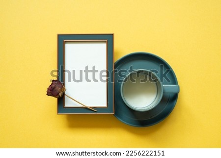 Empty navy photo frame and dry rose flower, cup of milk on yellow background. top view, copy space