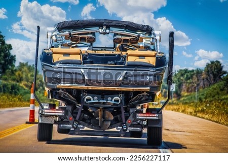 Taking the boat to the lake - Speedboat on boat trailer viewed from behind with blurred trees and foliage in background and construction cone to one side Royalty-Free Stock Photo #2256227117