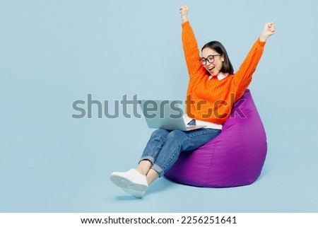 Full body happy young IT woman of Asian ethnicity wear orange sweater glasses sit in bag chair work hold use laptop pc computer do winner gesture isolated on plain pastel light blue cyan background