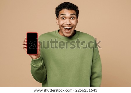 Young surprised man of African American ethnicity wearing green sweatshirt hold in hand use mobile cell phone with blank screen workspace area isolated on plain pastel light beige background studio