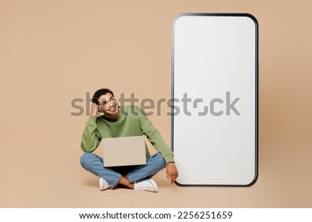 Full body young man of African American ethnicity wear green sweatshirt big huge blank screen mobile cell phone smartphone with area hold laptop pc computer isolated on plain pastel beige background