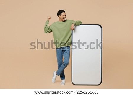 Full body young man of African American ethnicity wear green sweatshirt big huge blank screen mobile cell phone smartphone with area do winner gesture isolated on plain pastel light beige background