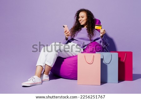 Full body young woman wear pullover sit in chair near package bags after shopping use mobile cell phone credit card isolated on plain pastel light purple background. Black Friday sale buy day concept