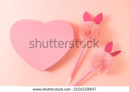 One heart-shaped gift box and two handles with a fluffy head and rabbit ears lies in the center on a natural beige stone background, flat lay close-up. Concept of valentine's day, happy easter.