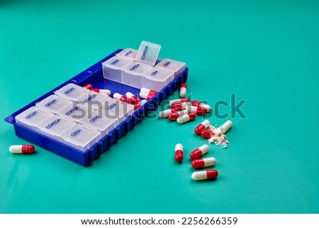 Pillbox of the days of the week on a green table with some red and white capsules on top. Photography in landscape format. Copy space.