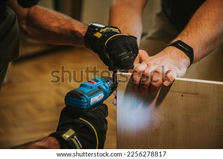 In the colorful horizontal detail image, handymen use a screwdriver to screw a screw into a piece of furniture in a naturally lit interior, which is carpentry work.