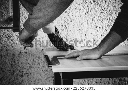 In a horizontal black and white detailed image, the handymen measure the thick furniture board with the tape measure and the protractor in an outdoor space with natural lighting.