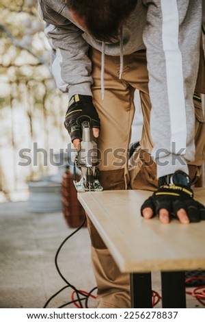 In a vertical color image, the handyman saws the wooden board with his saw in an outdoor space with natural light, which is a carpentry job.