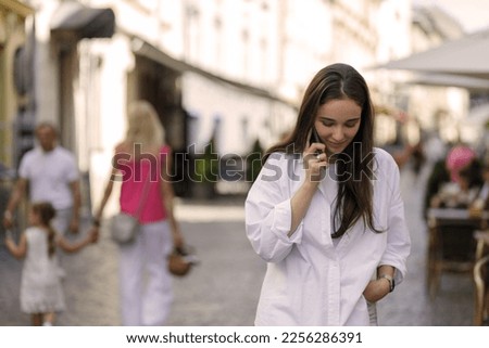 Young woman with dark long hair talking on phone walking through the street. Royalty-Free Stock Photo #2256286391