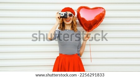 Summer portrait of happy smiling young woman with film camera and red heart shaped balloon wearing french beret hat on white background