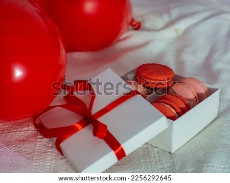 Opened gift box with macaron cookies on the bed. Gift box with sweets lying on white bed with a lot of red heart shape balloons. Copy space. Valentines Day. Buying gifts.