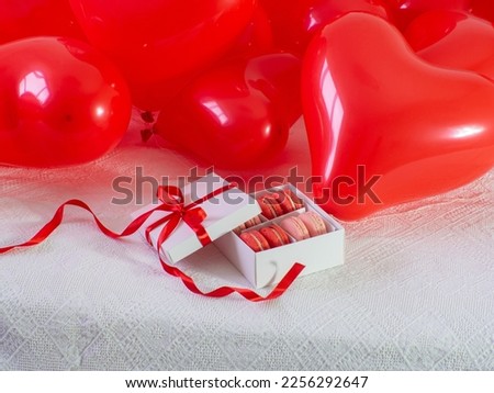 Opened gift box with macaron cookies on the bed. Gift box with sweets lying on white bed with a lot of red heart shape balloons. Copy space. Valentines Day. Buying gifts.