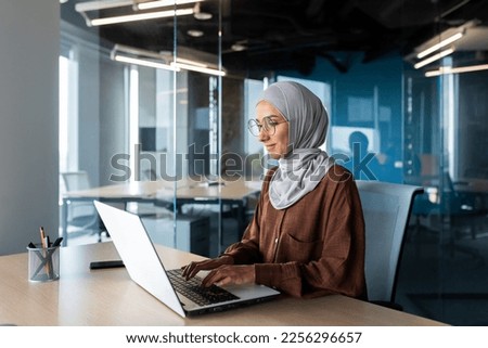Successful smiling Arab woman in hijab working inside modern office, Muslim woman using laptop at work, business woman satisfied with achievement results typing on computer keyboard. Royalty-Free Stock Photo #2256296657