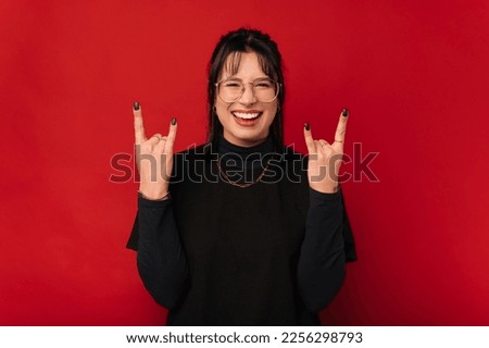 Cheerful young black haired woman is showing the rock gesture to the camera over red background.