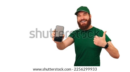 Cheerful delivery man in green shows the screen of his phone and a thumb up over white background.