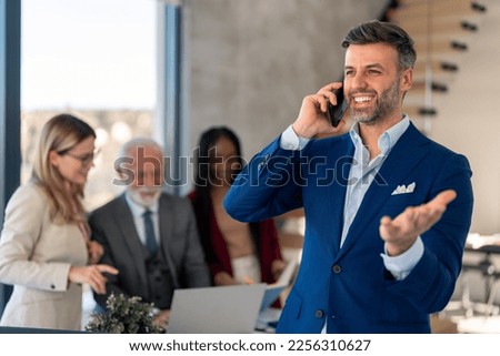 Trustworthy businessman in suit talking on phone making a deal with important client, working in modern office with multiethnic business team.