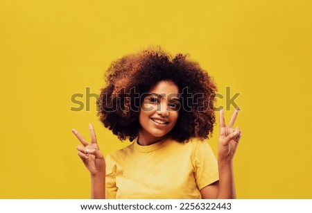 lifestyle, emotion and people concept: Cheerful laughing funny young african american woman standing showing victory sign looking camera isolated on yellow color wall background studio portrait