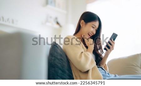 Happy young asian woman relax on comfortable couch at home texting messaging on smartphone, smiling girl use cellphone, chatting online message, shopping online from home Royalty-Free Stock Photo #2256345791