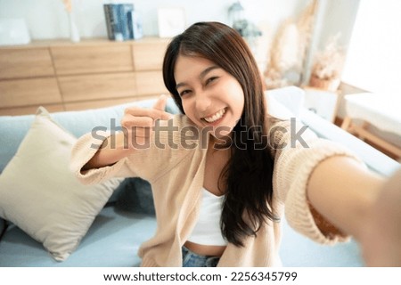 Pretty young asian female with big smile sitting at living room. She having fun taking light cheerful selfie on blurred background