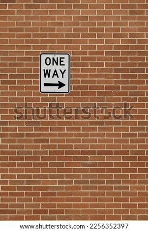 ONE WAY road sign with direction arrow pointing right. White road sign with black text and arrow icon. Sign is bolted to a red brick wall. Right pointing arrow, portrait orientation