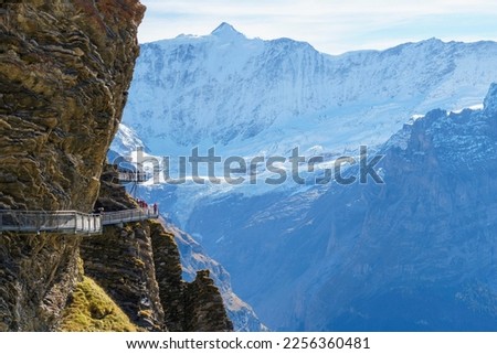 Tourists admire majestic snowy alpine mountains along a hair-raising sheer cliffwalk Royalty-Free Stock Photo #2256360481