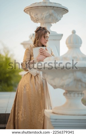 A beautiful young woman in an eighteenth century historical gold dress stands on the stairs of a mansion with a book in her hands. the princess is reading in the palace. fabulous image Royalty-Free Stock Photo #2256379341