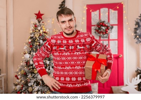 A young man in a red sweater in Scandinavian style holds a box with a gift in his hands. The guy on the background of the Christmas tree porch.