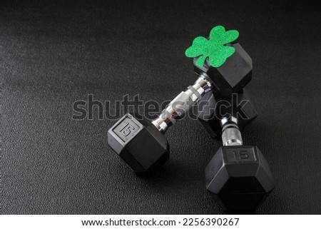 Set of hex head dumbbells with a green glitter shamrock on a black gym floor, happy St. Patrick’s Day
