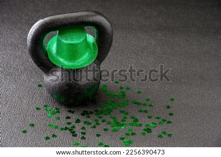 Black iron kettlebell, and green glitter leprechaun hat, covered in green shamrock confetti on a black gym floor, happy St. Patrick’s Day
