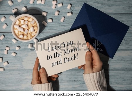 LOVE YOU TO THE MOON AND BACK text on valentine card inscription positive quote phrase. Female hands holding greeting card blue envelope with white cup of coffee and marshmallows on wooden blue