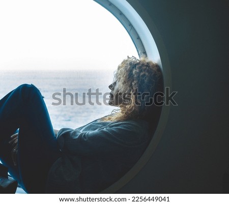 Serene woman sitting inside a porthole in the boat cruise ferry transport sea looking the ocean water and enjoying the trip alone. Passenger people dreaming destination. Relax and journey lifestyle Royalty-Free Stock Photo #2256449041