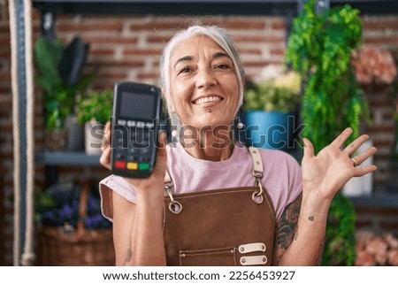 Middle age woman with tattoos working at florist shop holding dataphone celebrating achievement with happy smile and winner expression with raised hand 